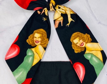 May include: A black headband with a red bow and a print of pin-up girls and red hearts. The headband is made of fabric and has a black elastic band.