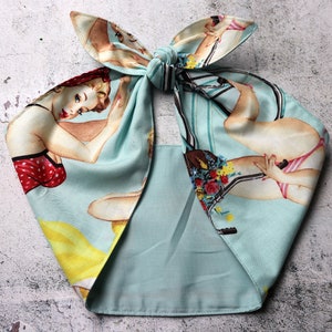 May include: A light blue headscarf with a vintage pin-up girl print. The fabric features illustrations of women in swimwear and bicycles. The scarf is tied in a bow at the top.