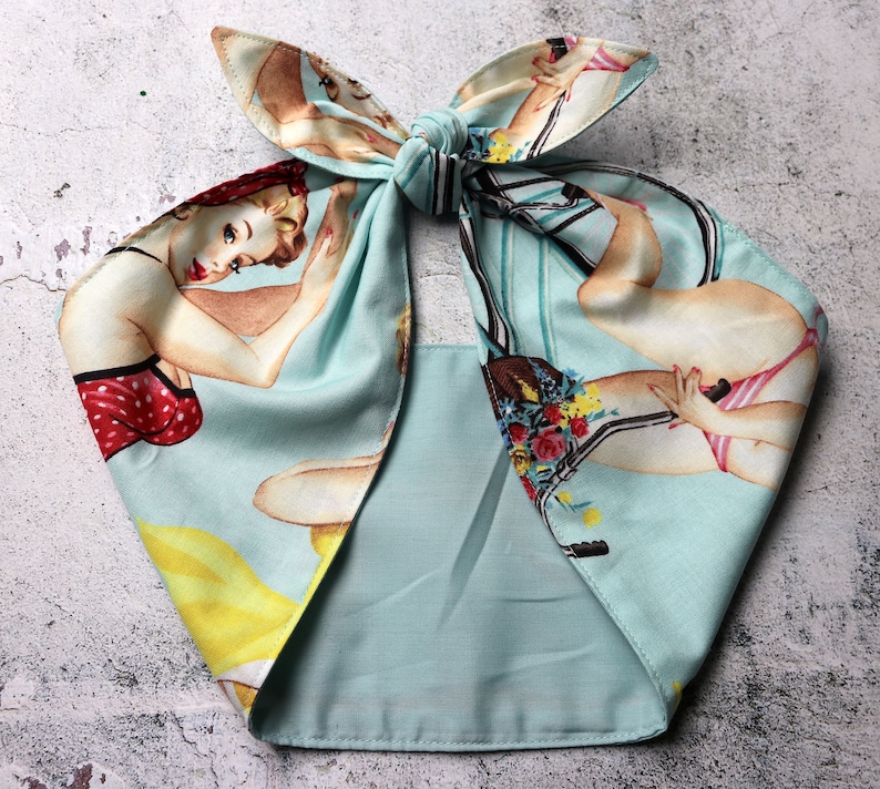 May include: A light blue headscarf with a vintage pin-up girl print. The fabric features illustrations of women in swimwear and bicycles. The scarf is tied in a bow at the top.