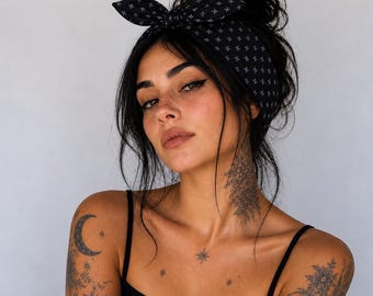 May include: A black headband with a bow, patterned with small white crosses, worn on the head. The headband is paired with a black tank top. The person has dark hair and tattoos on the arm and neck.