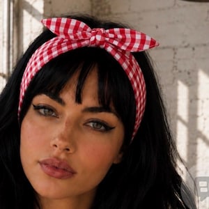 Puede incluir: Una diadema con estampado de cuadros vichy en rojo y blanco que presenta un detalle de lazo anudado en la parte superior. El accesorio de tela se coloca sobre la cabeza y muestra un diseño clásico con un nudo central sobre la frente.