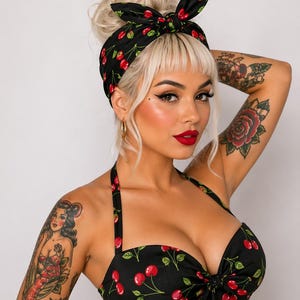 Puede incluir: Diadema negra y top halter con estampado de cerezas rojas. La diadema está atada con un lazo. El top tiene un escote corazón y un detalle de lazo. La mujer tiene el pelo rubio y lápiz labial rojo.