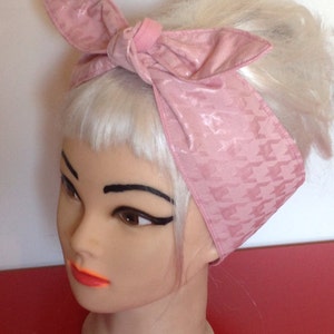 May include: A pink headband with a houndstooth pattern. The headband is tied in a bow at the top of the head.
