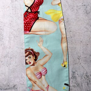 May include: A light blue fabric featuring vintage pin-up girls in swimwear. One woman is in a red polka dot swimsuit, another in a yellow dress, and a third in a pink and white striped bikini riding a bicycle with a flower basket.