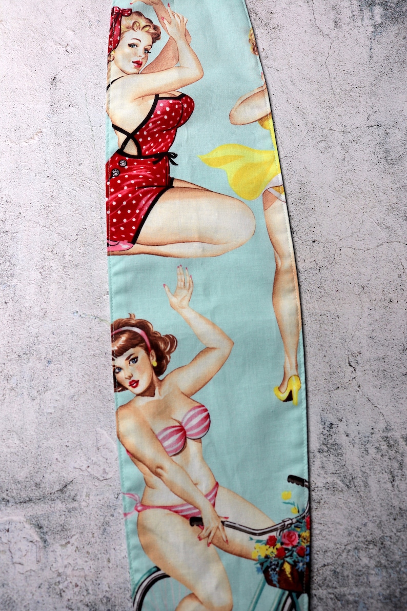 May include: A light blue fabric featuring vintage pin-up girls in swimwear. One woman is in a red polka dot swimsuit, another in a yellow dress, and a third in a pink and white striped bikini riding a bicycle with a flower basket.