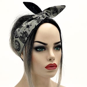 May include: A black and grey patterned headband with a bow. The headband displays a repeating pattern of occult symbols and illustrations. The bow is black with a similar design. The headband is worn on a mannequin head with dark hair.