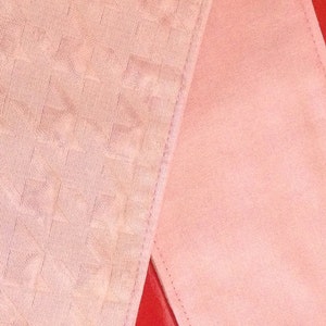 May include: Two pieces of pink fabric, one smooth and one textured, are layered on top of each other. The smooth fabric is on top and has a subtle sheen. The textured fabric has a repeating pattern of small squares.