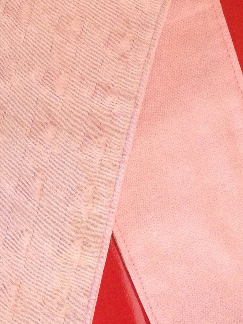 May include: Two pieces of pink fabric, one smooth and one textured, are layered on top of each other. The smooth fabric is on top and has a subtle sheen. The textured fabric has a repeating pattern of small squares.