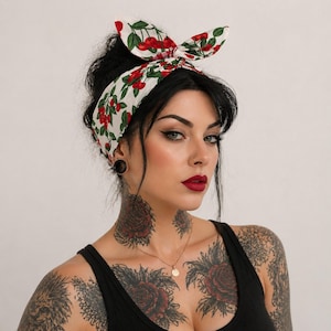 May include: A white fabric headband featuring a repeating pattern of red cherries and green leaves. The headband is tied at the top with a bow-like knot, showcasing a vibrant botanical print against a clean white background, designed for hair.