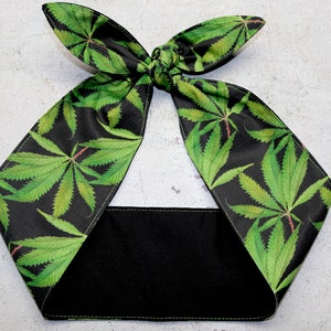 May include: A black headband featuring a pattern of green cannabis leaves. The headband is tied in a bow at the top and has a solid black band. White stitching details the edges of the fabric.