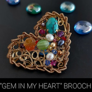 Puede incluir: Una broche de alambre en forma de corazón dorada con una variedad de gemas y cuentas de colores, incluyendo turquesa, naranja, verde y perlas blancas. La broche se titula "GEM IN MY HEART" BROOCH.