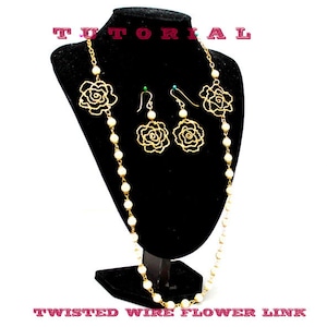 Puede incluir: Un collar de oro con cuentas de perlas blancas y detalles de alambre en forma de rosa. También se incluyen pendientes a juego con detalles de alambre en forma de rosa. El collar y los pendientes se muestran en un soporte negro. "Twisted Wire Flower Link" está escrito debajo del soporte.