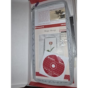 May include: A Husqvarna Viking Mega Hoop embroidery frame set in a white box. Includes a gray plastic hoop, a small clear container of metal clips, a set of clips in a plastic bag, an instruction sheet, and a red Mega Hoop Upgrade CD.