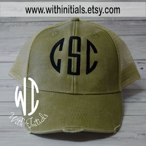 May include: A green distressed trucker hat with a black mesh back. The hat has a black embroidered monogram of the letters "CSC" in a circle.