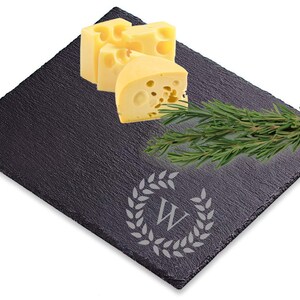 Puede incluir: Una tabla de queso de pizarra negra con una corona decorativa y la letra "W" grabada en el centro. La tabla est&aacute; cubierta con rebanadas de queso amarillo y una ramita de romero.