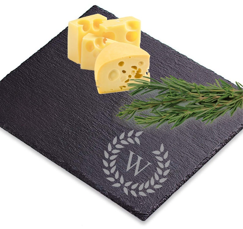 Puede incluir: Una tabla de queso de pizarra negra con una corona decorativa y la letra "W" grabada en el centro. La tabla est&aacute; cubierta con rebanadas de queso amarillo y una ramita de romero.