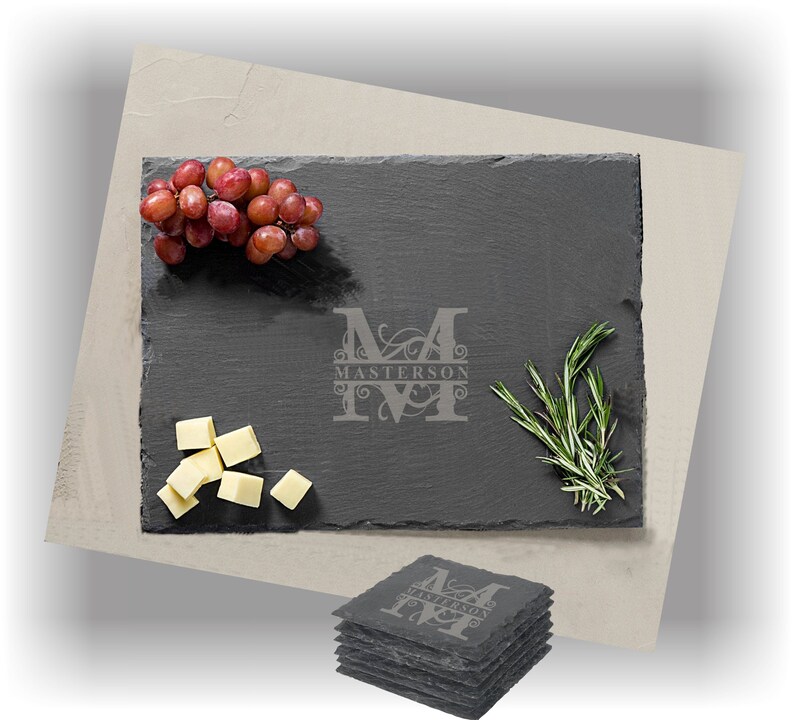 Puede incluir: Una tabla de servir de pizarra negra con una inicial "M" monograma y el nombre "Masterson" grabado en una escritura decorativa. La tabla est&aacute; rodeada de uvas, cubos de queso y ramitas de romero.