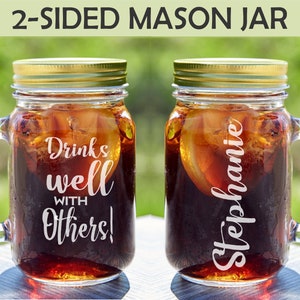May include: Two clear glass mason jars with handles, filled with a dark beverage and ice. One jar has the text "Drinks well WITH Others!" and the other has the name "Stephanie". Both jars have gold lids.