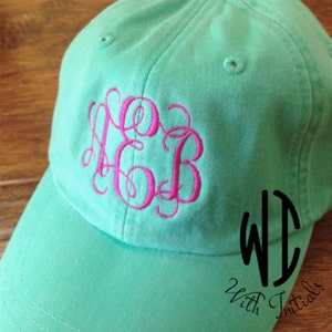 May include: A turquoise blue baseball cap with a pink monogram embroidered on the front. The monogram is in a script font and reads "KGB". The cap also has a black logo on the side that reads "With Initials".