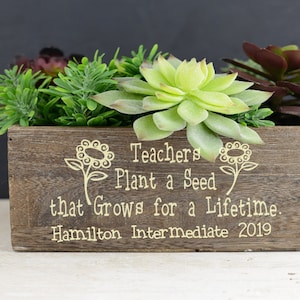 May include: A wooden planter box with a succulent arrangement. The planter is engraved with the text "Teachers Plant a Seed that Grows for a Lifetime. Hamilton Intermediate 2019".