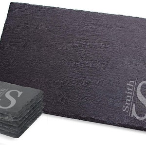 Puede incluir: Un juego de seis posavasos de pizarra negra con una inicial y un nombre personalizados, "Smith S", grabados en color plateado. Los posavasos est&aacute;n apilados uno encima del otro.