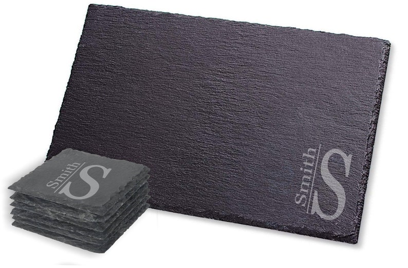 Puede incluir: Un juego de seis posavasos de pizarra negra con una inicial y un nombre personalizados, "Smith S", grabados en color plateado. Los posavasos est&aacute;n apilados uno encima del otro.