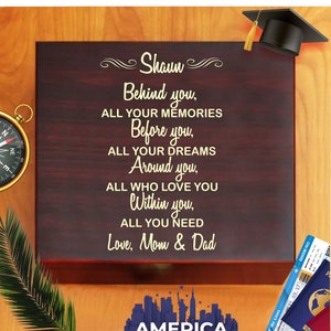 May include: A dark wood box with a gold inscription that reads "Shaun Behind you, ALL YOUR MEMORIES Before you, ALL YOUR DREAMS Around you, ALL WHO LOVE YOU Within you, ALL YOU NEED Love, Mom & Dad". The box is decorated with a small compass and a graduation cap. The box is sitting on a wooden surface with a cityscape and the word "AMERICA" printed below.