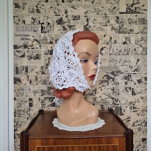 Può includere: Un cappello bianco all'uncinetto con un motivo a pizzo. Il cappello è su una testa di manichino con capelli rossi. Il manichino è su un tavolo di legno con un pizzo bianco.