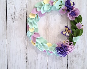 May include: A pastel green, yellow and purple felt wreath with flowers and a butterfly. The wreath is made of felt leaves and flowers.