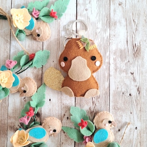 May include: A brown felt platypus keyring with a gold glitter tail, wearing a flower crown. The platypus is surrounded by felt flowers and leaves.