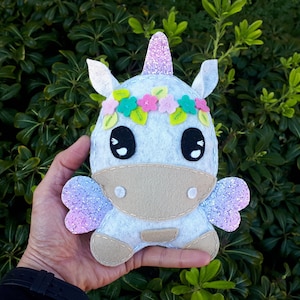 May include: A plush unicorn toy with a white body, pink and purple glitter wings, and a pink horn. The unicorn has a flower crown with pink, white, and purple flowers.