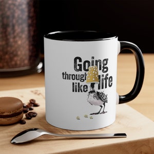 Op de afbeelding: Witte keramische koffiemok met een zwarte rand en handvat. De mok heeft een zwart-witte illustratie van een vogel die een papieren beker op zijn kop draagt. De tekst "Going through life like" staat op de mok gedrukt.