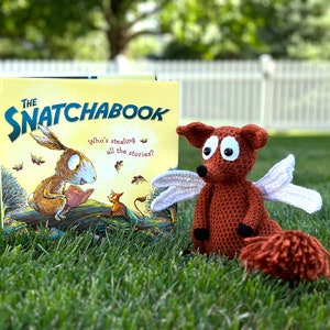 May include: A crocheted fox with wings sitting on green grass next to a book titled "The Snatchbook: Who's stealing all the stories?"