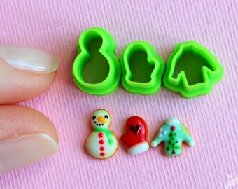 Op de afbeelding: Groene plastic koekjesvormen in de vorm van een sneeuwpop, want en trui, naast drie gedecoreerde suikerkoekjes: een sneeuwpop, een want en een trui.