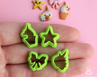 May include: A collection of colorful polymer clay charms and green plastic cookie cutters on a pink surface. The charms include a yellow star, a bow, a unicorn, and a cupcake. The cookie cutters are shaped like a unicorn, a star, and a bow.