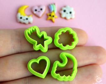 May include: Green plastic cookie cutters in the shapes of a star, cloud, heart, and crescent moon. The cutters are on a hand against a pink background.