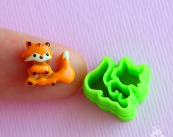 May include: A small orange fox figurine with white accents and brown eyes. The figurine is sitting on a finger. Next to the fox is a green plastic mold shaped like a bush.