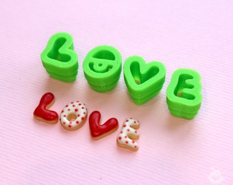 May include: Green plastic cookie cutters in the shape of the letters "LOVE" and a heart.  The cutters are stacked on top of each other.  There are also red and white cookies in the shape of the letters "LOVE" on a pink surface.