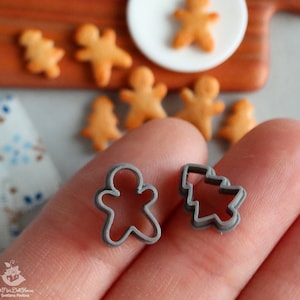 Könnte beinhalten: Zwei graue Metall-Ausstecher, einer in Form eines Lebkuchenmanns und der andere in Form eines Weihnachtsbaums. Die Ausstecher werden in der Hand einer Person gehalten.