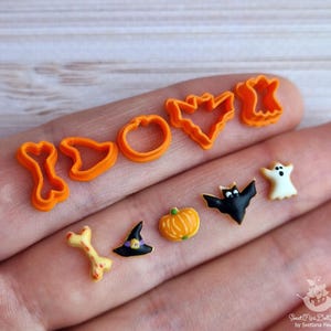 Può includere: Un set di nove tagliapasta di Halloween in plastica arancione a forma di zucca, cappello da strega, pipistrello, fantasma, osso e fantasma. I tagliapasta sono sulla mano di una persona.