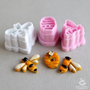May include: A set of three pink and white plastic cookie cutters shaped like bees and honeycombs. There are also three decorated cookies, two bees and one honeycomb.