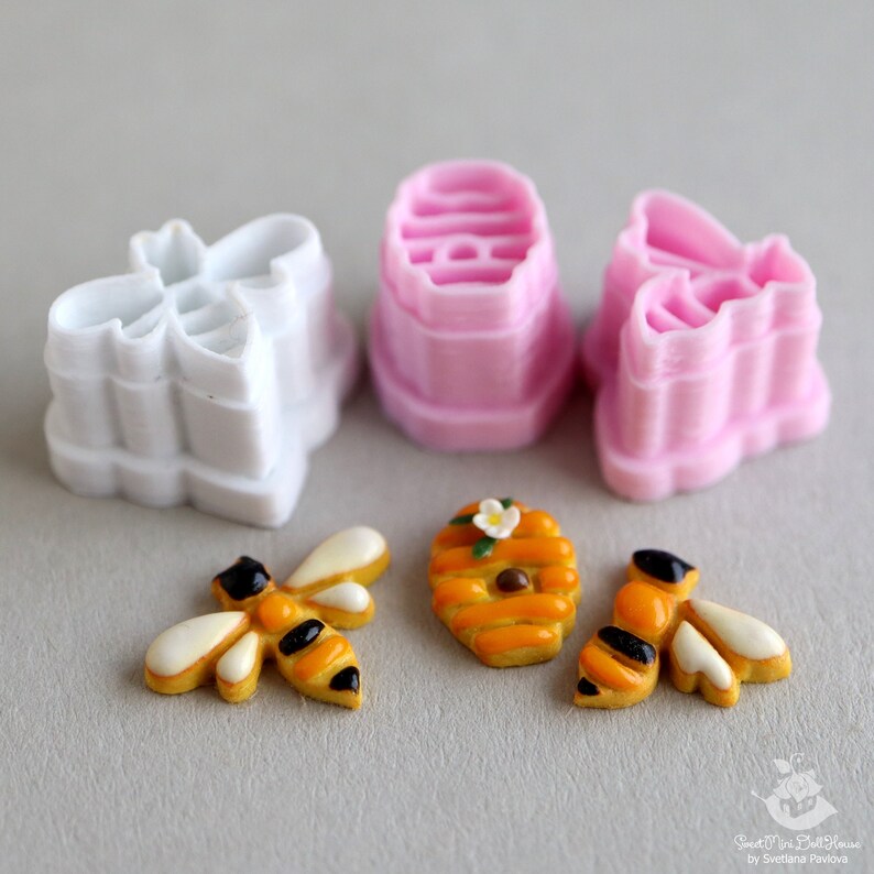 May include: A set of three pink and white plastic cookie cutters shaped like bees and honeycombs. There are also three decorated cookies, two bees and one honeycomb.
