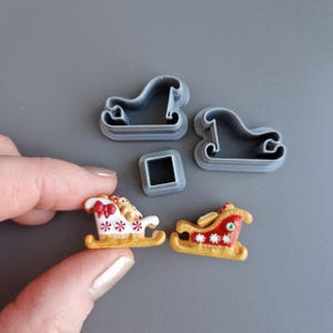 Puede incluir: Primer plano de cortadores de galletas y galletas decoradas con temática navideña. Los cortadores son de plástico gris con forma de trineo y cuadrado. Dos galletas decoradas tienen forma de trineo, una con lazo rojo y otra con estampado rojo.