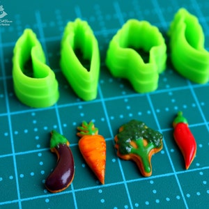 May include: Green plastic cookie cutters shaped like leaves, carrots, an eggplant, a broccoli, and a chili pepper. The cutters are on a green cutting mat with white dots.