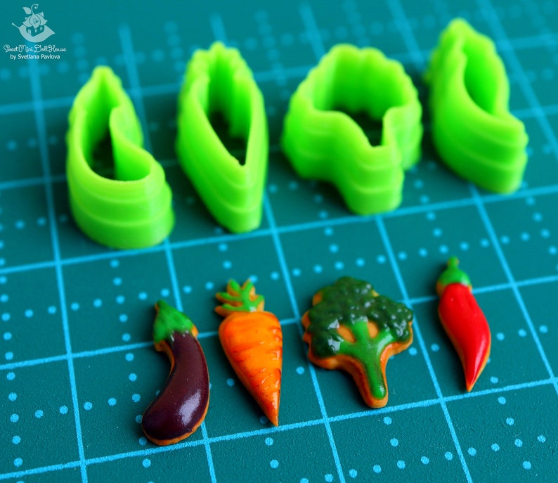 May include: Green plastic cookie cutters shaped like leaves, carrots, an eggplant, a broccoli, and a chili pepper. The cutters are on a green cutting mat with white dots.