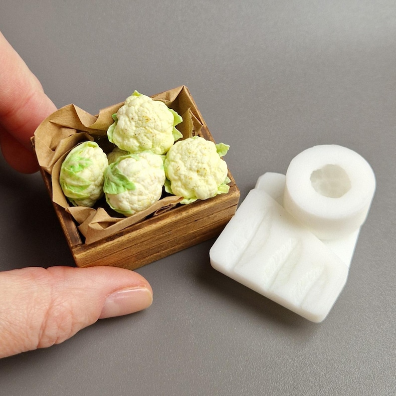 Pu&ograve; includere: Piccola cassa di legno piena di mini cavolfiori dettagliati. La cassa &egrave; foderata con carta marrone. Accanto c'&egrave; un oggetto bianco rettangolare con una parte superiore circolare. Miniature per casa delle bambole o progetto artigianale.