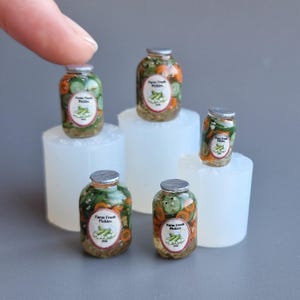 Op de afbeelding: Miniatuur glazen potten gevuld met augurken, komkommers en wortelen. Elke pot heeft een zilveren deksel en een etiket met de tekst "Farm Fresh Pickles". De potten zijn gerangschikt op witte platforms.