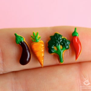 May include: Four miniature food items made of polymer clay: a green eggplant, an orange carrot, a green broccoli, and a red chili pepper.