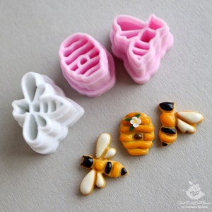 May include: A set of bee-themed cookie cutters and fondant decorations. The set includes two bee-shaped cutters, one honeycomb cutter, and two fondant bees with a honeycomb design. The cutters are white plastic and the fondant decorations are yellow and black.