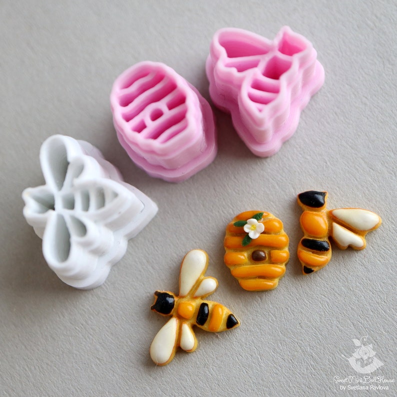 May include: A set of bee-themed cookie cutters and fondant decorations. The set includes two bee-shaped cutters, one honeycomb cutter, and two fondant bees with a honeycomb design. The cutters are white plastic and the fondant decorations are yellow and black.